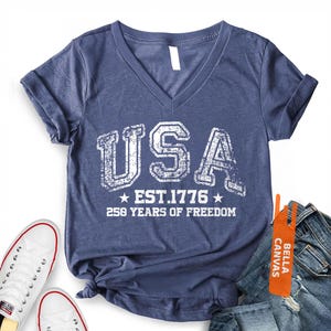 Peut inclure: T-shirt bleu marine chin&eacute; avec col en V, orn&eacute; du mot "USA" en lettres blanches us&eacute;es, "EST. 1776" et "250 YEARS OF FREEDOM". Le t-shirt est pr&eacute;sent&eacute; avec des baskets blanches et un jean bleu.