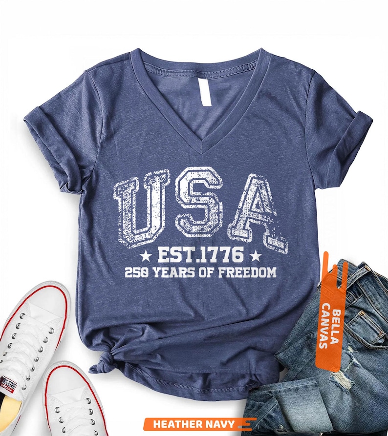 Peut inclure: T-shirt bleu marine chin&eacute; avec col en V, orn&eacute; du mot "USA" en lettres blanches us&eacute;es, "EST. 1776" et "250 YEARS OF FREEDOM". Le t-shirt est pr&eacute;sent&eacute; avec des baskets blanches et un jean bleu.