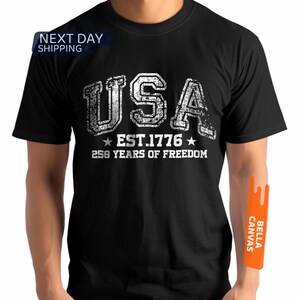 Peut inclure: T-shirt noir avec un motif blanc vieilli indiquant "USA EST. 1776 250 YEARS OF FREEDOM". Le t-shirt a un col rond et semble &ecirc;tre en coton.