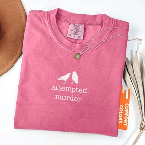 May include: A pink t-shirt with the words "attempted murder" and two bird illustrations. The shirt has a crew neck and a tag that says "Comfort Colors". A silver necklace with a pendant is on the shirt.
