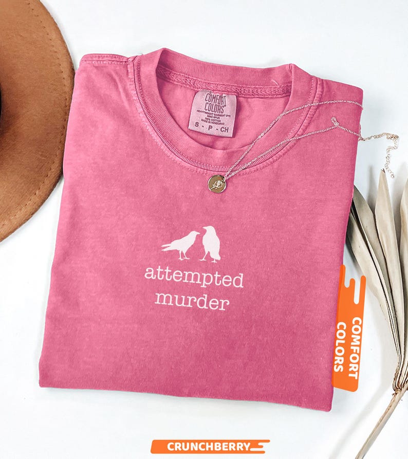 May include: A pink t-shirt with the words "attempted murder" and two bird illustrations. The shirt has a crew neck and a tag that says "Comfort Colors". A silver necklace with a pendant is on the shirt.