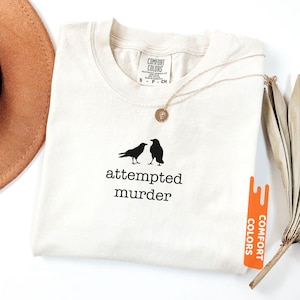 May include: Ivory t-shirt featuring the words "attempted murder" and two black bird silhouettes. A gold necklace with a pendant lies on the shirt. The shirt has a Comfort Colors label.
