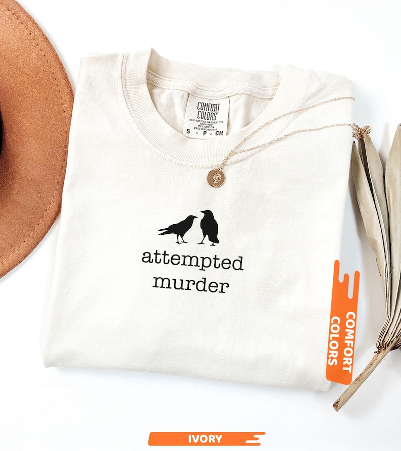 May include: Ivory t-shirt featuring the words "attempted murder" and two black bird silhouettes. A gold necklace with a pendant lies on the shirt. The shirt has a Comfort Colors label.