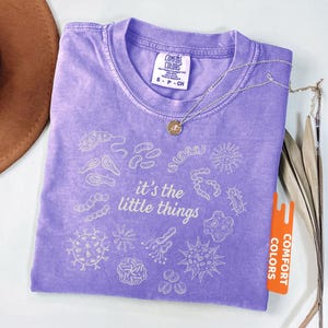 May include: A violet Comfort Colors t-shirt with the phrase "it's the little things" and various microbe illustrations. A silver necklace with a small pendant rests on the shirt. The shirt has a small tag that says "Comfort Colors".
