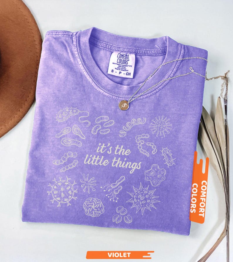 May include: A violet Comfort Colors t-shirt with the phrase "it's the little things" and various microbe illustrations. A silver necklace with a small pendant rests on the shirt. The shirt has a small tag that says "Comfort Colors".