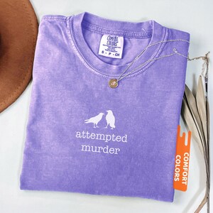 May include: A violet Comfort Colors t-shirt featuring the words "attempted murder" and a graphic of two birds. A silver necklace with a pendant is on the shirt. The shirt has a small tag that says "Comfort Colors".