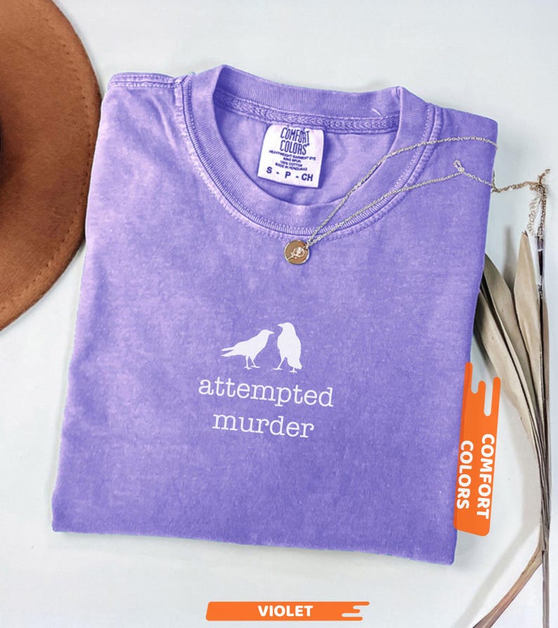 May include: A violet Comfort Colors t-shirt featuring the words "attempted murder" and a graphic of two birds. A silver necklace with a pendant is on the shirt. The shirt has a small tag that says "Comfort Colors".