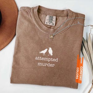 May include: Espresso-coloured Comfort Colors t-shirt with the words "attempted murder" and two bird silhouettes in white. A silver necklace with a pendant rests on the shirt. The shirt is folded and next to a brown hat.