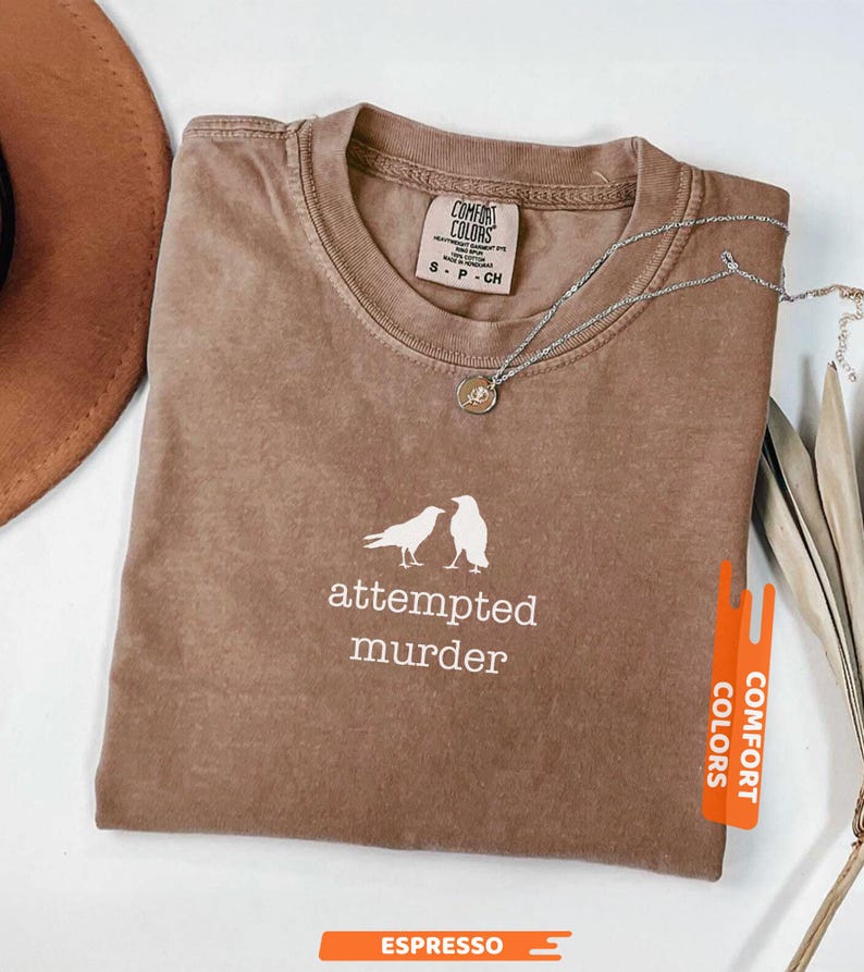 May include: Espresso-coloured Comfort Colors t-shirt with the words "attempted murder" and two bird silhouettes in white. A silver necklace with a pendant rests on the shirt. The shirt is folded and next to a brown hat.