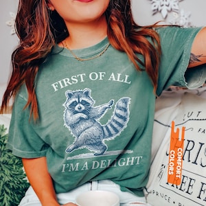 Puede incluir: Camiseta verde claro con el texto "FIRST OF ALL I'M A DELIGHT" y un gr&aacute;fico de mapache azul. La camiseta es de la marca Comfort Colors. La camiseta es de color verde claro.
