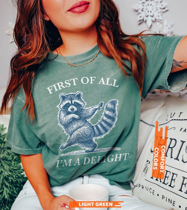 Puede incluir: Camiseta verde claro con el texto "FIRST OF ALL I'M A DELIGHT" y un gr&aacute;fico de mapache azul. La camiseta es de la marca Comfort Colors. La camiseta es de color verde claro.
