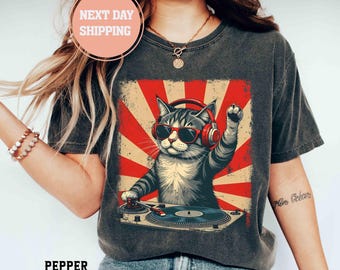 May include: A charcoal gray t-shirt featuring a graphic of a tabby cat wearing red headphones and sunglasses, DJing on a turntable. The background of the graphic consists of red and cream-colored sunburst rays. The shirt has a distressed texture.