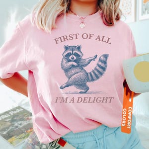 Puede incluir: Camiseta rosa claro con un dibujo de mapache y el texto "FIRST OF ALL I'M A DELIGHT". El mapache est&aacute; representado bailando. La camiseta tiene mangas cortas y cuello redondo.