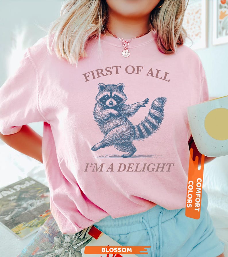 Puede incluir: Camiseta rosa claro con un dibujo de mapache y el texto "FIRST OF ALL I'M A DELIGHT". El mapache est&aacute; representado bailando. La camiseta tiene mangas cortas y cuello redondo.