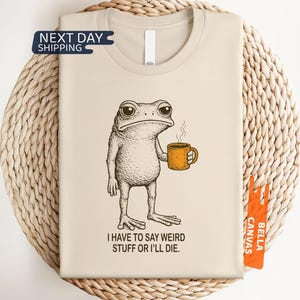 May include: A natural-colored t-shirt featuring a black and white line art illustration of a frog holding an orange mug with steam rising from it. Below the frog, the text reads, I HAVE TO SAY WEIRD STUFF OR I'LL DIE. The shirt has a crew neckline.