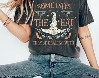 May include: A charcoal gray short-sleeve t-shirt featuring a graphic of a witch hat with spiderwebs and the text: SOME DAYS YOU JUST HAVE TO PUT IN THE HAT AND REMIND THEM WHO THEY'RE DEALING WITH. The shirt is paired with blue denim jeans.