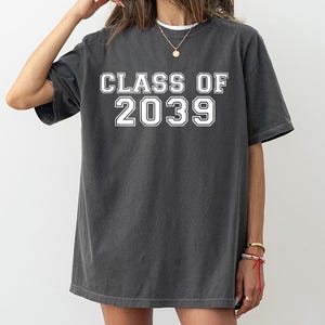 May include: A charcoal gray short-sleeve t-shirt featuring the white text "CLASS OF 2039" printed in a bold, collegiate-style font across the chest. The garment has a crew neckline and a relaxed, oversized fit with a slightly faded fabric finish.