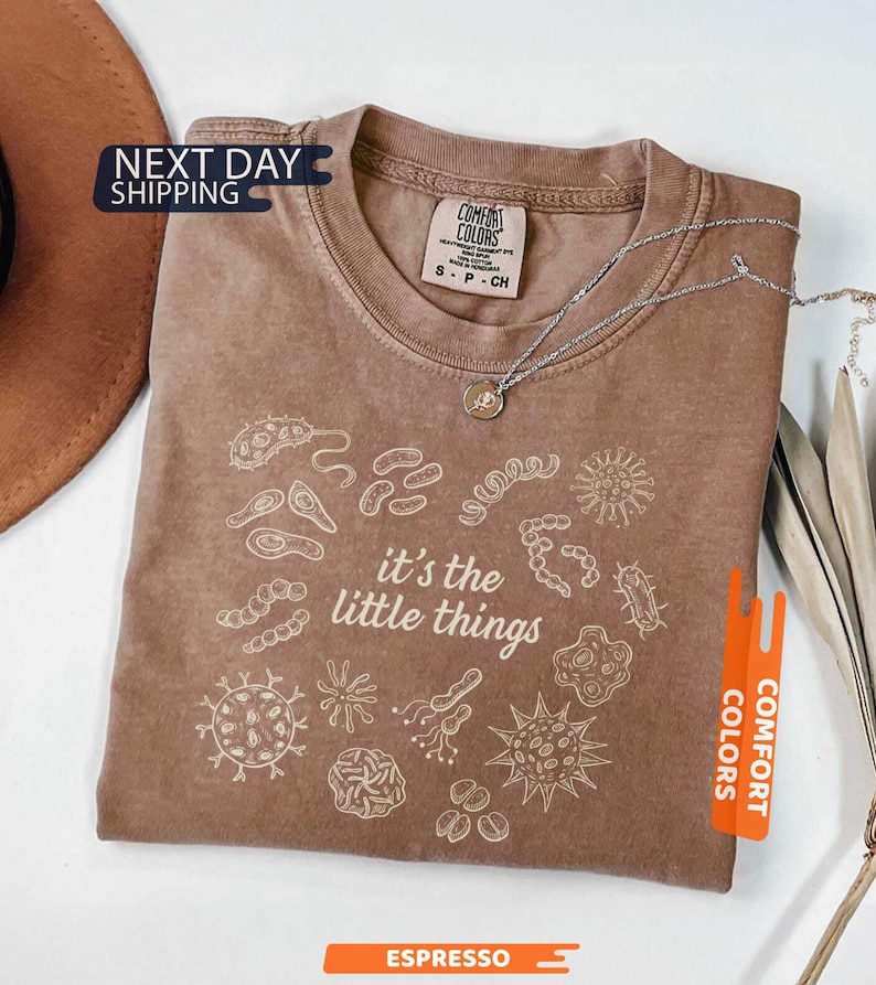 May include: Espresso-colored Comfort Colors t-shirt with the phrase "it's the little things" and various microbe illustrations in white. A silver necklace with a small pendant rests on the shirt. The shirt is folded and displayed on a white surface.