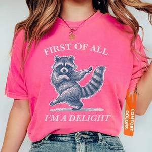 Puede incluir: Una camiseta rosa intenso con el texto "FIRST OF ALL I'M A DELIGHT" y un dibujo de un mapache. El mapache est&aacute; representado en una pose de baile. La camiseta tiene una etiqueta Comfort Colors.