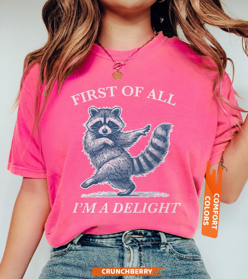 Puede incluir: Una camiseta rosa intenso con el texto "FIRST OF ALL I'M A DELIGHT" y un dibujo de un mapache. El mapache est&aacute; representado en una pose de baile. La camiseta tiene una etiqueta Comfort Colors.