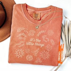 May include: Coral-colored t-shirt with white illustrations of microorganisms and the phrase "it's the little things." A gold necklace with a pendant rests on the shirt. The shirt has a tag that says "Comfort Colors."
