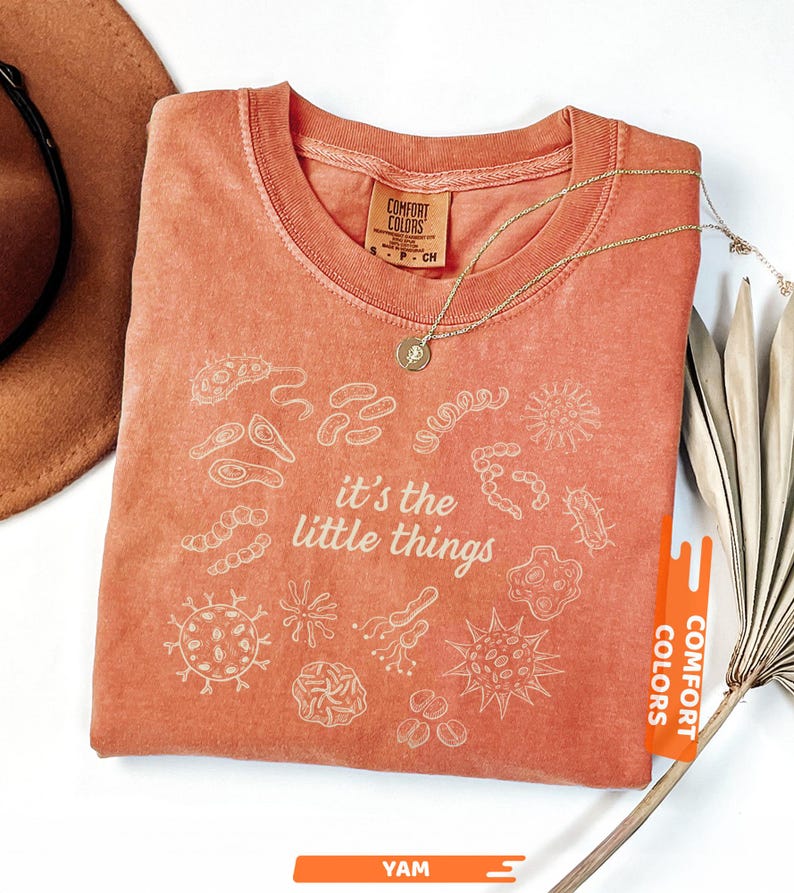 May include: Coral-colored t-shirt with white illustrations of microorganisms and the phrase "it's the little things." A gold necklace with a pendant rests on the shirt. The shirt has a tag that says "Comfort Colors."