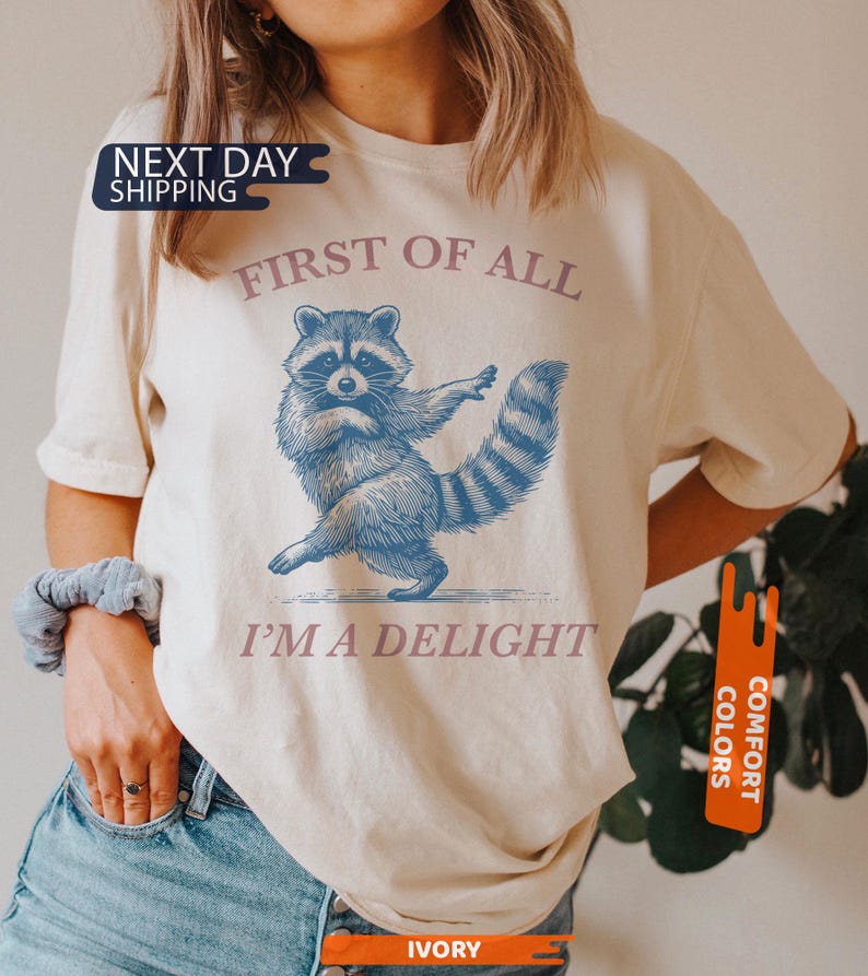 Puede incluir: Camiseta color marfil con una ilustraci&oacute;n de mapache azul y el texto "FIRST OF ALL I'M A DELIGHT". El mapache est&aacute; representado bailando. La camiseta es de la marca Comfort Colors.