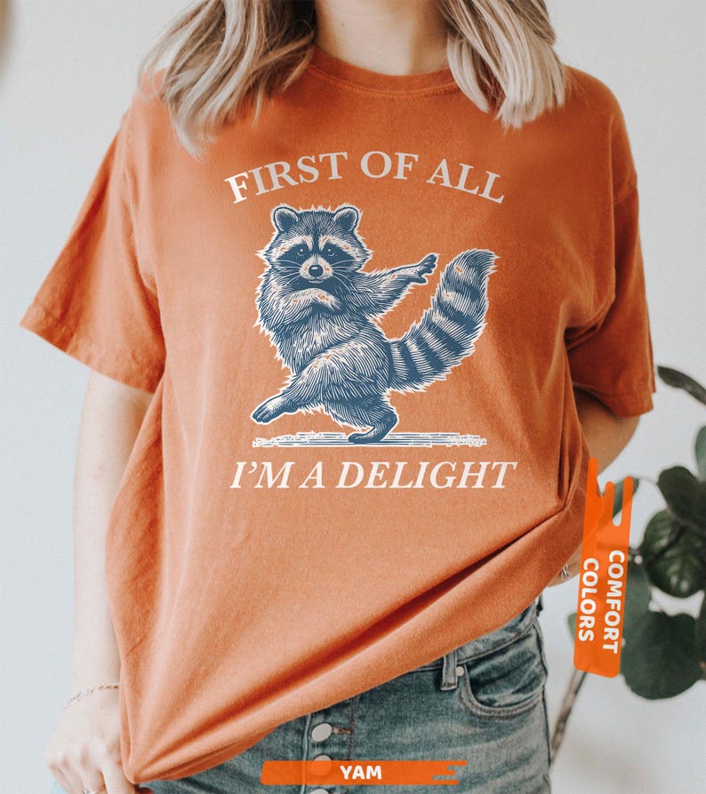 Puede incluir: Camiseta naranja quemado con un gr&aacute;fico de mapache azul y blanco. El texto "FIRST OF ALL I'M A DELIGHT" est&aacute; impreso en blanco. La camiseta tiene mangas cortas y un corte holgado. La etiqueta de la camiseta dice "COMFORT COLORS".
