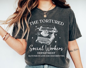 May include: A charcoal gray short-sleeve t-shirt featuring a white graphic of a vintage typewriter. The text reads THE TORTURED Social Workers DEPARTMENT ALL'S FAIR IN LOVE AND DOCUMENTING. The design includes small star accents surrounding the typewriter.