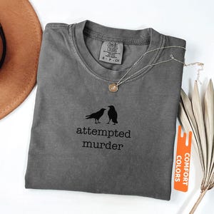 May include: Grey t-shirt with the words "attempted murder" and a graphic of two crows. The shirt has a crew neck and short sleeves. A silver necklace with a pendant is draped on the shirt. The shirt is labelled "Comfort Colors".
