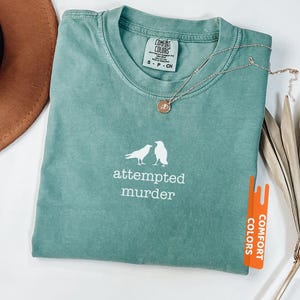 May include: A light green Comfort Colors t-shirt with the words "attempted murder" and a graphic of two birds. A gold necklace with a small pendant rests on the shirt. The shirt is labelled "Light Green".