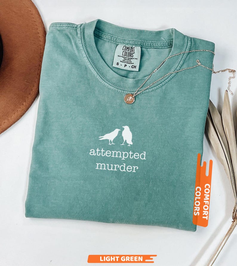May include: A light green Comfort Colors t-shirt with the words "attempted murder" and a graphic of two birds. A gold necklace with a small pendant rests on the shirt. The shirt is labelled "Light Green".