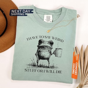 May include: A sage green t-shirt featuring a black ink-style illustration of a frog holding a coffee mug. The text above the frog reads I HAVE TO SAY WEIRD and the text below reads STUFF OR I WILL DIE. The shirt has a Comfort Colors brand label.