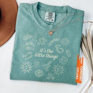 May include: Light green t-shirt with the phrase "it's the little things" and various microbe illustrations. The shirt has a crew neck and short sleeves. A silver necklace with a small pendant is draped across the shirt.