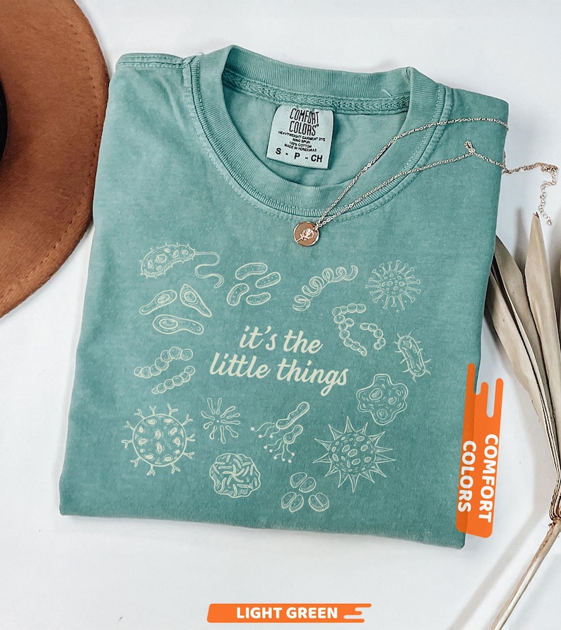 May include: Light green t-shirt with the phrase "it's the little things" and various microbe illustrations. The shirt has a crew neck and short sleeves. A silver necklace with a small pendant is draped across the shirt.