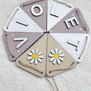 May include: A wooden banner spelling "LOVE" in white, attached to triangular pieces in shades of white, beige, and mauve. Two triangles feature daisy cutouts. Each piece is connected with twine for hanging.