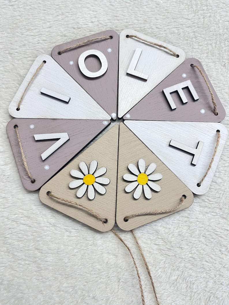 May include: A wooden banner spelling "LOVE" in white, attached to triangular pieces in shades of white, beige, and mauve. Two triangles feature daisy cutouts. Each piece is connected with twine for hanging.