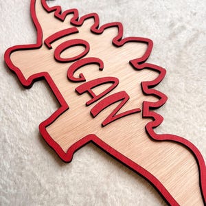 May include: Wooden dinosaur-shaped sign with a red outline and the name "LOGAN" written in red cursive lettering.
