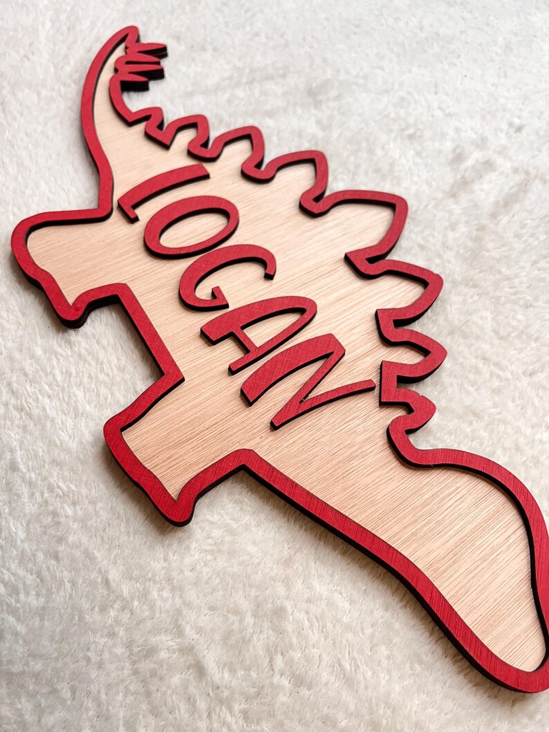 May include: Wooden dinosaur-shaped sign with a red outline and the name "LOGAN" written in red cursive lettering.