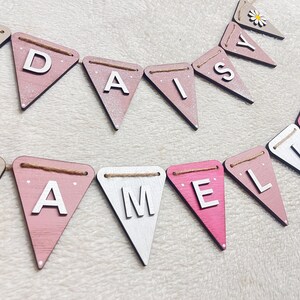 May include: Wooden bunting spelling out "DAISY" and "AMELIA" in white capital letters on pink, white, and beige pennants. Each pennant is strung with twine and some feature a daisy design. A charming, personalised decoration.