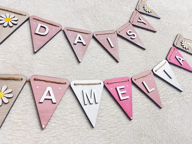 May include: Wooden bunting spelling out "DAISY" and "AMELIA" in white capital letters on pink, white, and beige pennants. Each pennant is strung with twine and some feature a daisy design. A charming, personalised decoration.