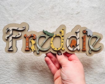 May include: Wooden name sign with the name "Freddie" spelled out in individual letters. Each letter is decorated with a different animal print and a small animal cutout. The letters are painted in shades of brown, yellow, orange, and green.