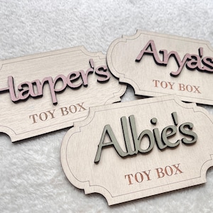 May include: Three wooden toy box labels with rounded edges. The labels are painted white and have the names "Harper's", "Anya's", and "Albie's" written in pink, brown, and green lettering, respectively. Each label also has the words "Toy Box" written in brown lettering.
