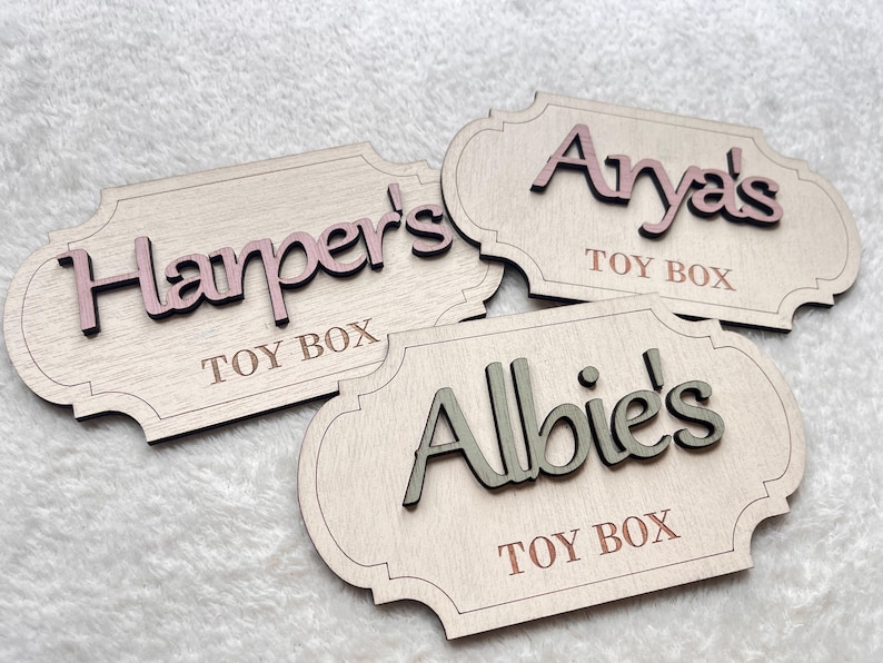 May include: Three wooden toy box labels with rounded edges. The labels are painted white and have the names "Harper's", "Anya's", and "Albie's" written in pink, brown, and green lettering, respectively. Each label also has the words "Toy Box" written in brown lettering.