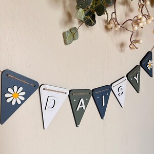 May include: A wooden banner spelling "DAISY" with pennant flags in blue, white, and green. One flag features a daisy design. The banner is strung on twine and is a decorative item, suitable for a nursery or home decor.