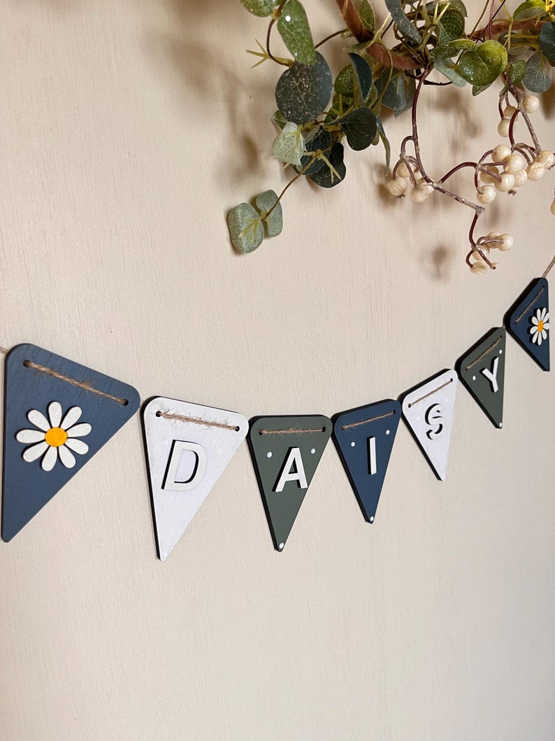 May include: A wooden banner spelling "DAISY" with pennant flags in blue, white, and green. One flag features a daisy design. The banner is strung on twine and is a decorative item, suitable for a nursery or home decor.