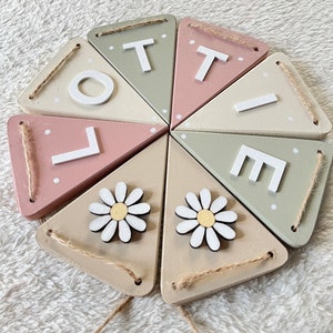 May include: A wooden banner spelling "LOTTE" in white letters, with daisy accents. The banner features triangular flags in pastel colours: pink, green, and beige. Each flag is connected with twine.