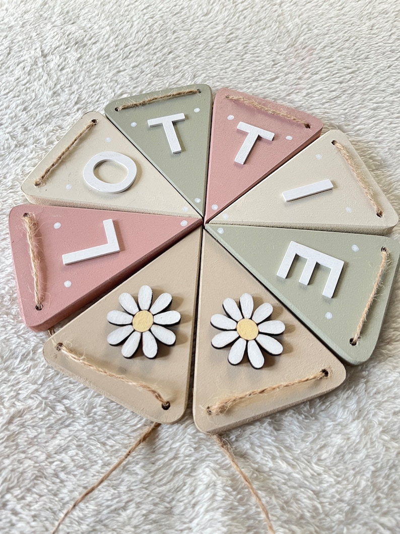 May include: A wooden banner spelling "LOTTE" in white letters, with daisy accents. The banner features triangular flags in pastel colours: pink, green, and beige. Each flag is connected with twine.