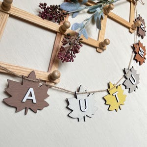 Könnte beinhalten: Eine dekorative Girlande mit Ahornblatt-Ausschnitten aus Holz in Braun-, Grau- und Gelbtönen, die jeweils einen weißen Buchstaben tragen, um das Wort AUTUMN zu bilden. Die Blätter sind an einer dünnen Schnur befestigt und hängen an einem Holzregal.