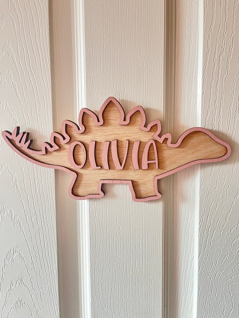 May include: A wooden dinosaur cutout with the name "Olivia" written inside. The dinosaur is light pink and has a brown wood grain texture.